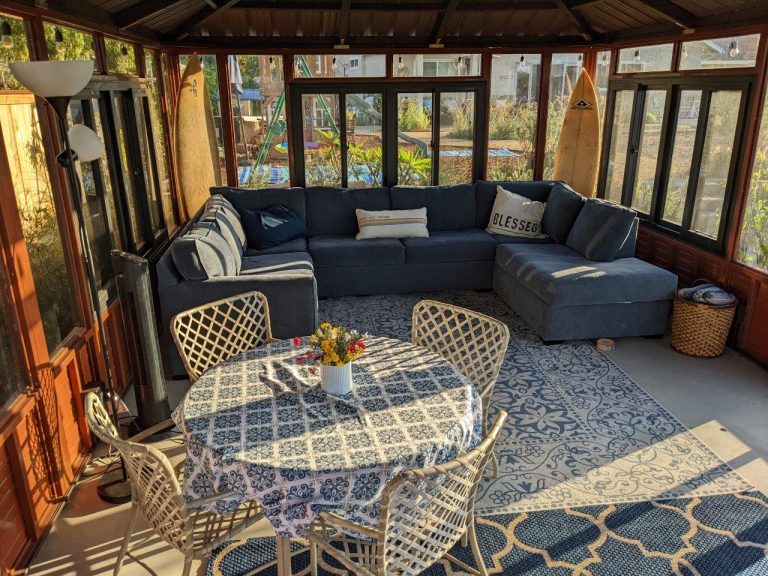 Sunlit sunroom with a blue sectional sofa, patterned rug, round table with floral tablecloth, and wicker chairs.