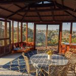 Sunlit wooden patio with round table, chairs, patterned rug, kids' play area, and large windows with an outdoor view.