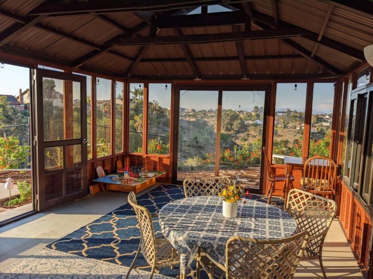 Sunlit wooden patio with round table, chairs, patterned rug, kids' play area, and large windows with an outdoor view.
