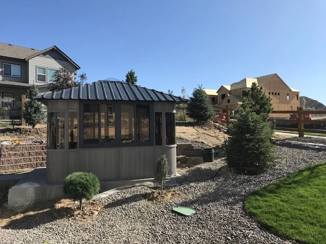 Small, modern gazebo in a landscaped yard with trees and gravel, houses and construction visible in the background.