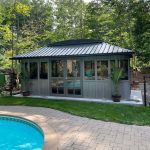 A garden gazebo with large windows, surrounded by trees, sits beside a pool on a sunny day.