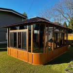 Wooden gazebo with large windows, set on grass in a fenced backyard under clear blue sky.
