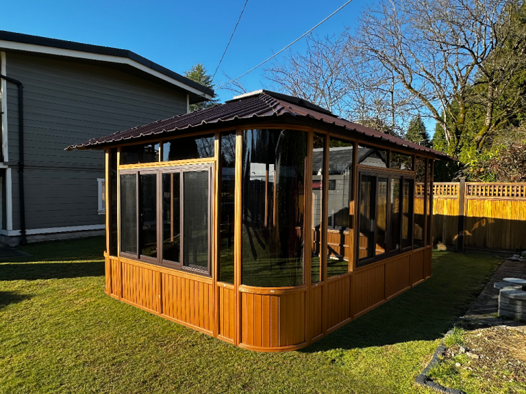 Wooden gazebo with large windows, set on grass in a fenced backyard under clear blue sky.