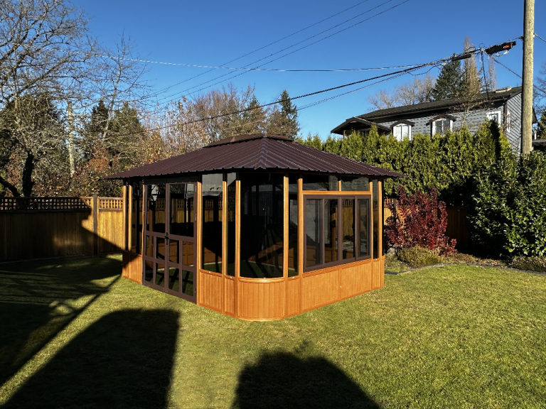 Wooden gazebo with glass windows on a sunny lawn, surrounded by a wooden fence and trees with a clear blue sky.