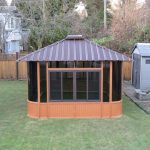 Wooden gazebo with a metal roof in a fenced backyard, with a shed to the right and trees in the background.