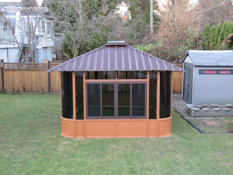 Wooden gazebo with a metal roof in a fenced backyard, with a shed to the right and trees in the background.