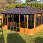 Wooden gazebo with glass windows, metal roof, in a backyard surrounded by fence and greenery.