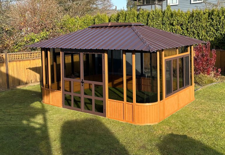 Wooden gazebo with glass windows, metal roof, in a backyard surrounded by fence and greenery.