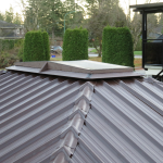 Brown metal roof with a small rectangular skylight, surrounded by trees and residential houses in the background.