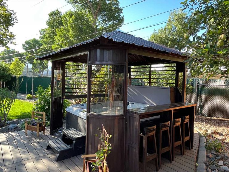 A wooden gazebo with a metal roof, enclosing a hot tub and bar seating, set in a lush, green backyard.
