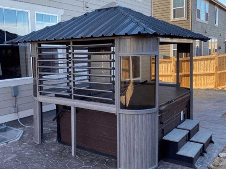 Outdoor gazebo covering a hot tub, with gray slatted walls and a metal roof, next to a house and wooden fence.