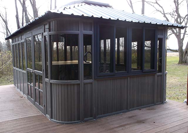 Rectangular gazebo with dark metal frame and large windows on a wooden deck, surrounded by leafless trees.