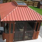 The Aspen 12x16 gazebo, featuring a red metal roof, large windows, and a skylight, is on sale. It sits on a concrete patio in a fenced backyard with grass and a wooden shed in the background.