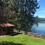 Lakeside scene with a small gazebo, trees, wooden fence, and mountains under a clear blue sky.