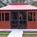 An Aspen 12x16 wooden gazebo with large windows and a reddish metal roof sits on a concrete foundation in a grassy yard, surrounded by trees and neighboring houses.