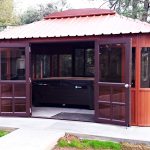 The Aspen 12x16, a wooden gazebo with a red metal roof on a concrete pad, features screened doors and windows. Inside, a large hot tub makes it perfect as a swim spa enclosure nestled among trees and grass.