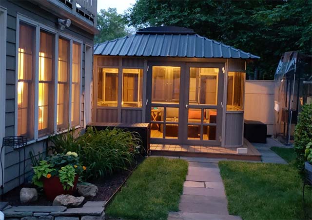 Lit gazebo with glass doors next to a house at dusk, surrounded by a garden and pathway.