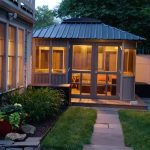 Cozy lit gazebo next to a house, surrounded by a garden path and greenery at dusk.