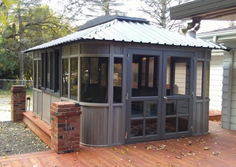 Small wooden gazebo with glass windows, metal roof, and red brick accents, set on a wooden deck surrounded by trees.