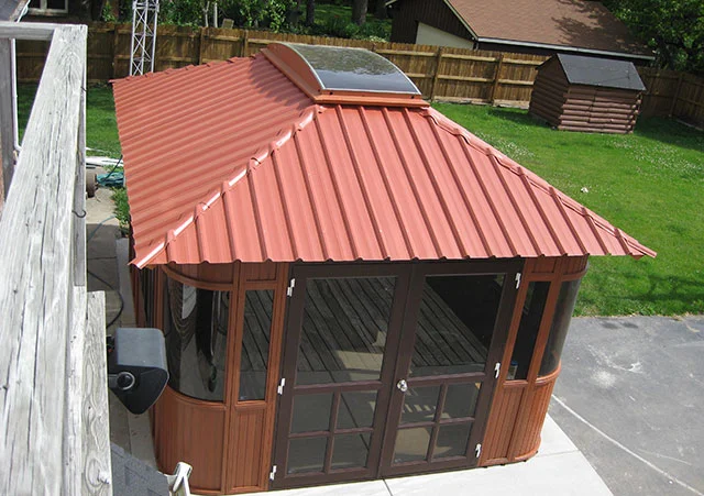 Red-roofed gazebo with glass doors and windows, set in a yard with green grass and wooden fences.