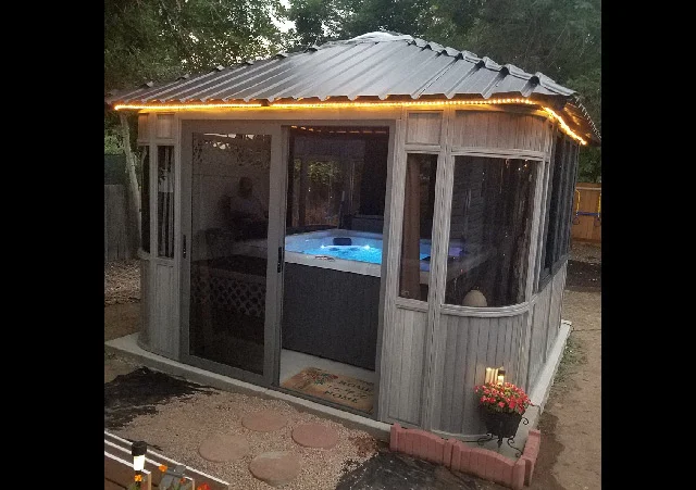 Gazebo with illuminated roof and glowing hot tub inside, surrounded by trees and garden elements.