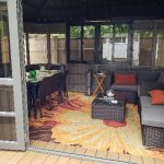 Enclosed wooden gazebo with dining table, chairs, and a cushioned sofa set on a colorful floral rug.