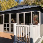 Gray gazebo with white railings on a wooden deck, surrounded by autumn trees under a clear blue sky.