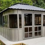 Gray metal garden shed with glass windows and doors, set on a concrete base, surrounded by greenery.