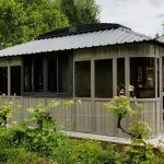 Small gray metal greenhouse with dark windows, surrounded by lush greenery, next to a residential building.