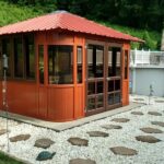 Red gazebo with windows, surrounded by white gravel and stepping stones, adjacent to a swimming pool and lush greenery.