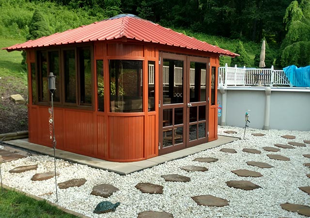 Red gazebo with windows, surrounded by white gravel and stepping stones, adjacent to a swimming pool and lush greenery.