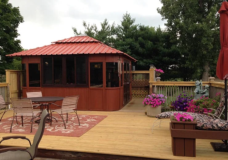 Wooden deck with a red-roofed gazebo, outdoor furniture, potted flowers, and trees in the background.