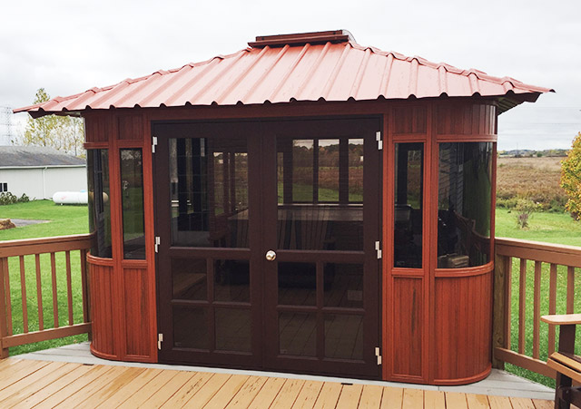 Wooden gazebo with brown trim and a red metal roof on a deck, set in a grassy backyard.