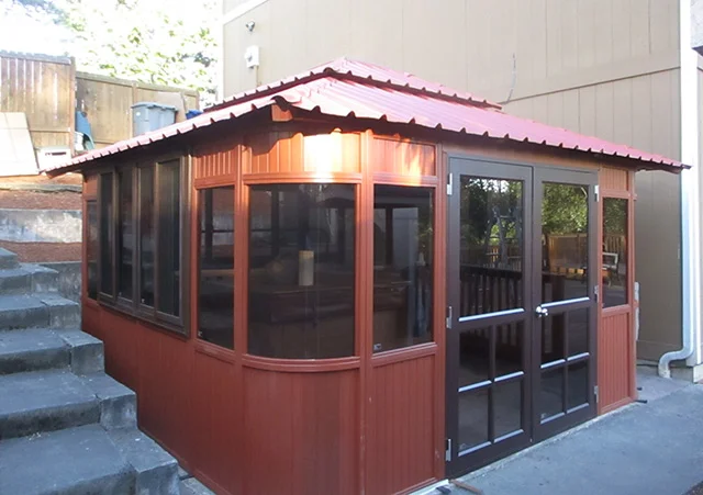 Red wooden gazebo with glass windows and doors, under a corrugated metal roof beside concrete steps.