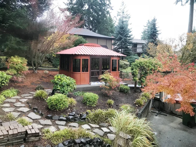 A garden with a red-roofed gazebo surrounded by trees and shrubs, featuring a stone pathway and grassy plants.