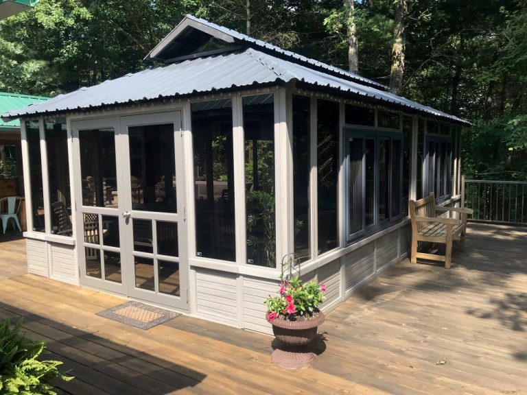 A screened gazebo with a metal roof on a wooden deck, surrounded by trees, with a bench and potted flowers.