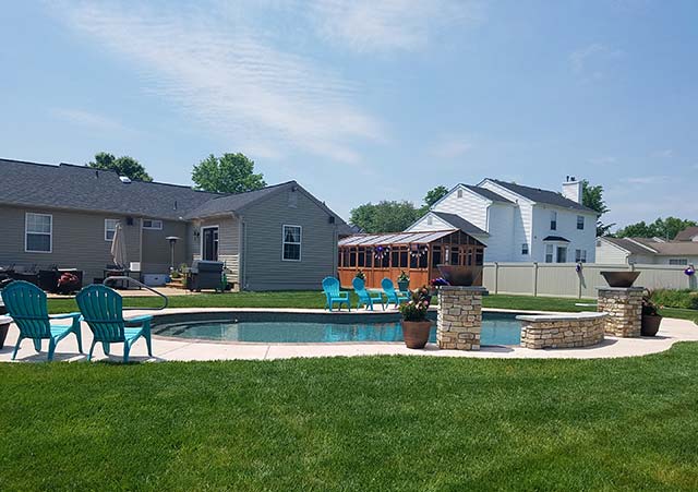 Backyard with a pool, patio chairs, and gazebo, surrounded by houses and green lawn under a clear blue sky.