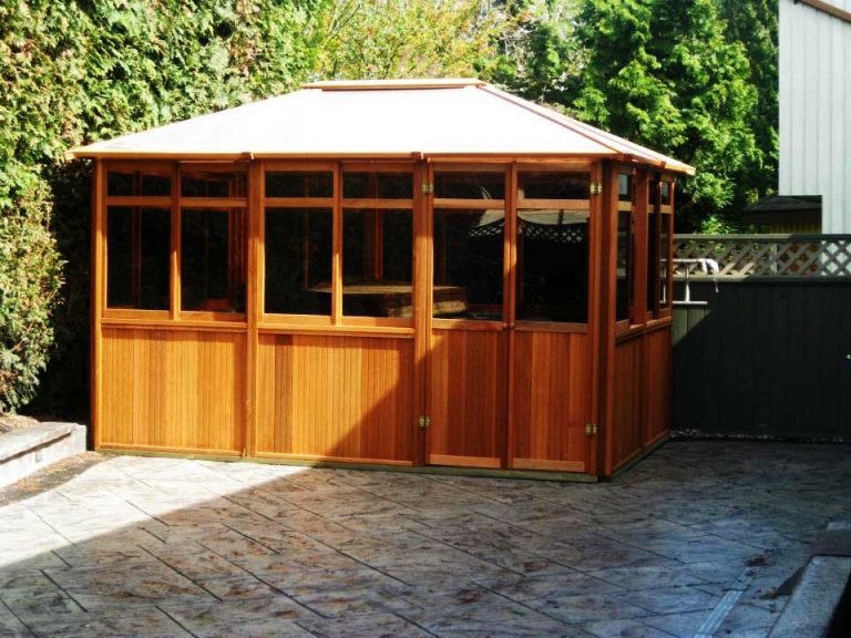 Wooden gazebo with large windows on a stone patio, surrounded by greenery.