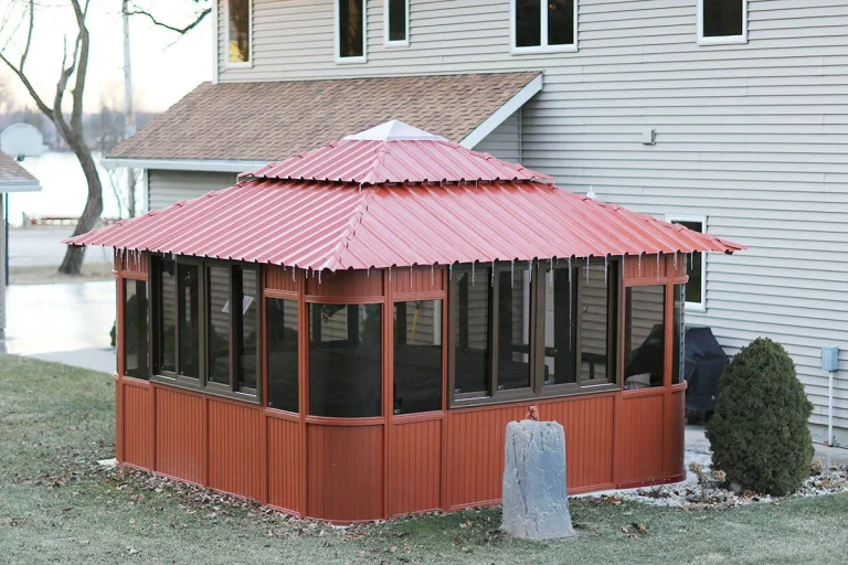 Red metal-roofed gazebo with glass panels, next to a gray house and small tree.