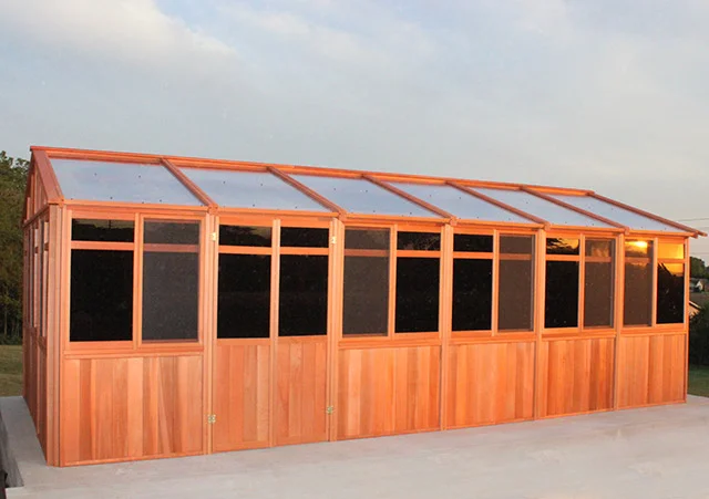 Wooden greenhouse with clear roof panels and dark windows, set on a concrete base against a sunset sky.