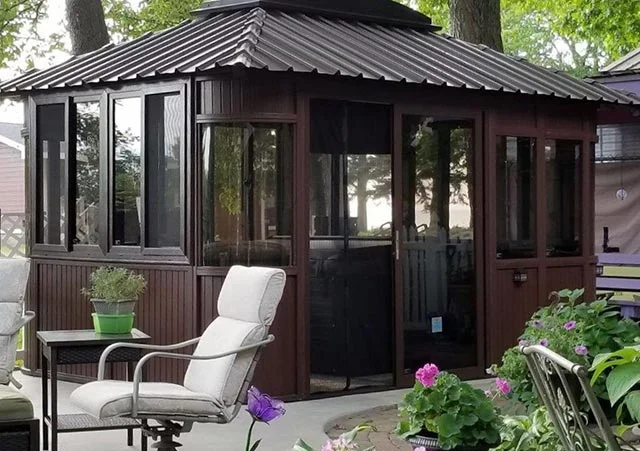 Brown gazebo with large windows, metal roof, surrounded by chairs and purple flowers on a patio.