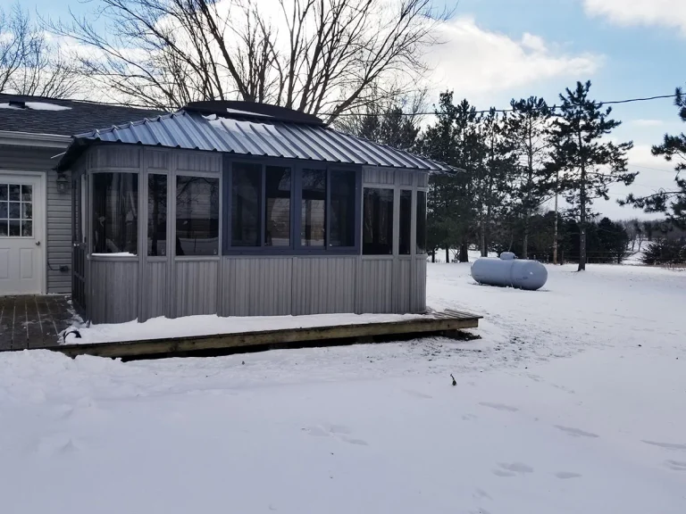 A snow-covered sunroom attached to a house, with trees and a propane tank in the background.