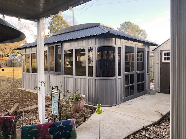 Modern metal gazebo with large windows and a dark roof on a concrete patio in a backyard.