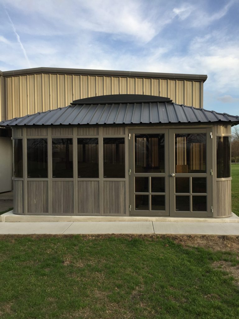 Small pavilion with metal roof and glass windows, set on a concrete slab, with grass in the foreground.