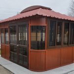 A gazebo with a red metal roof and glass windows, set on a concrete pad in a backyard.