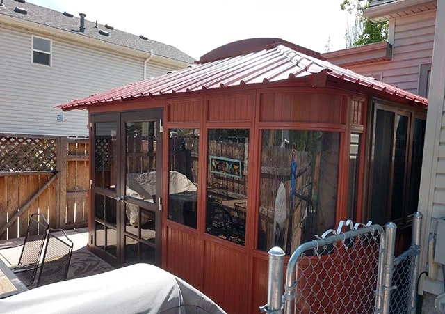 Red wooden gazebo with glass windows on a patio, surrounded by wooden fencing and neighboring houses.