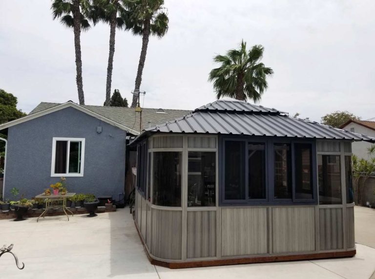 House with a gray gazebo on concrete, palm trees in the background, and potted plants on a table.