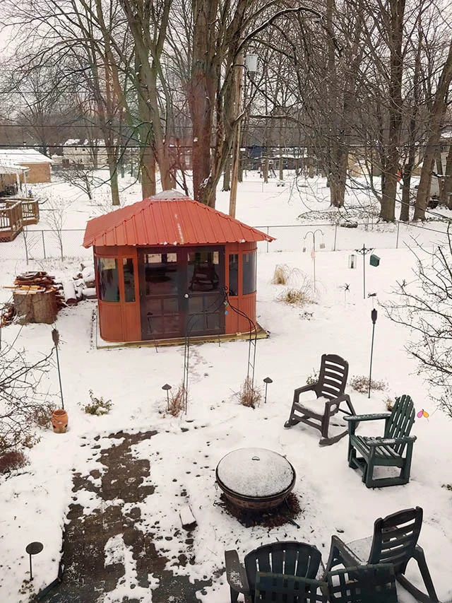 Snowy backyard with a red gazebo, patio chairs, and a fire pit surrounded by trees.
