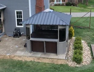 Backyard view of a small gazebo attached to a house, set on a stone patio with nearby landscaping and green grass.