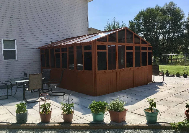 Wooden sunroom attached to a house, surrounded by patio furniture and potted plants on a sunny day.
